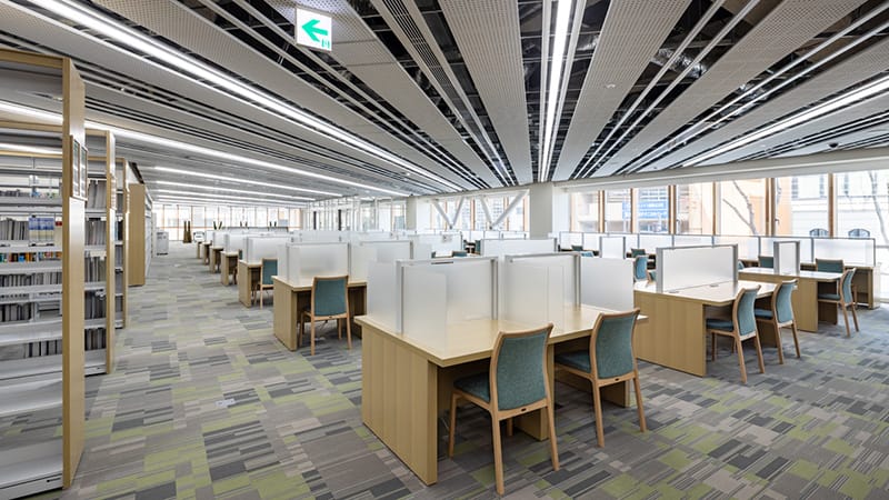 駅前キャンパスの図書室内、明るい窓際に並ぶ学習スペースと椅子が見える。