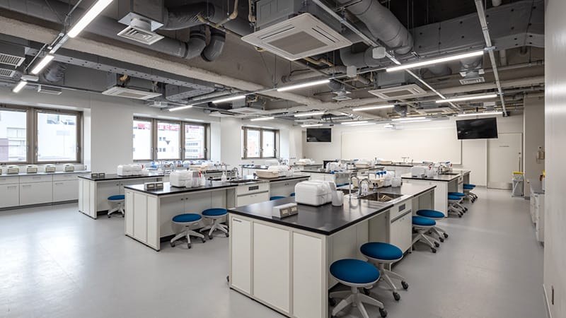 分析化学検査実習室の内部、白い作業台と青い椅子が並ぶ明るい空間。