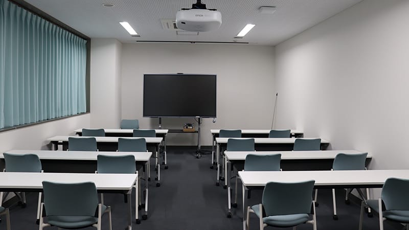 院生講義室1の内部、白い壁と青いカーテン、机と椅子が整然と配置されている。