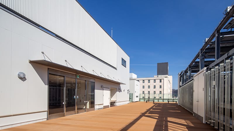 駅前キャンパスの屋上、明るい青空の下に広がる木製デッキと近代的な建物の風景。