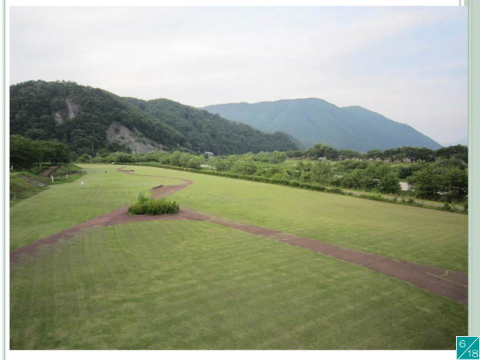 広々とした緑の公園と背後にそびえる山々の風景。晴れた空の下、自然が広がる静かな場所。