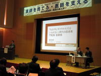 福島医学会のシンポジウムで講演を行う講師と聴衆の様子、背景にはテーマが掲示されたスクリーンがある。