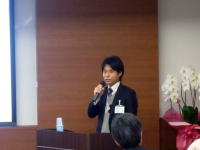 福島県立医科大学の講演会で話す男性がマイクを持ち、聴衆に向けてプレゼンテーションを行っている様子。