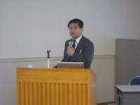 福島県立医科大学で講演を行う男性が、スライドを前にして話している様子。