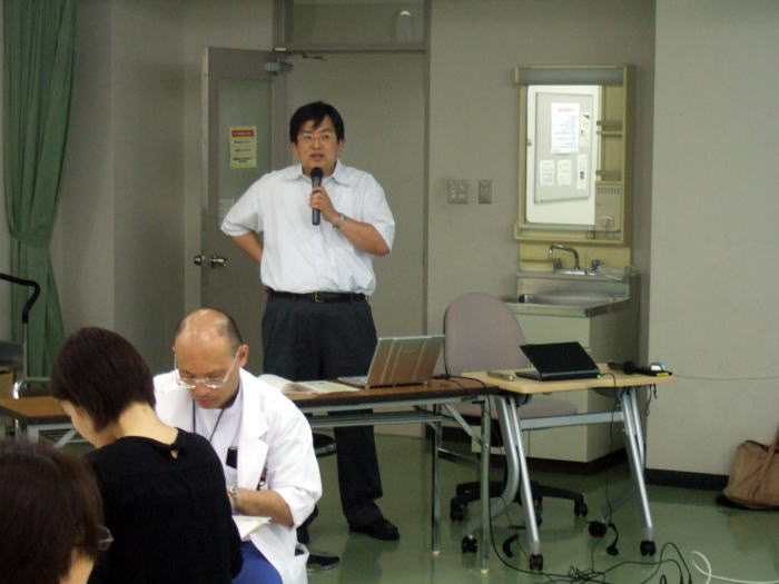 セミナーで講演を行う男性と、参加者が座っている様子。背景には机や椅子が配置されている。