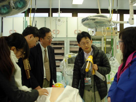 医療シミュレーション室での説明を受ける訪問者たち。医療機器や人形が配置された環境。