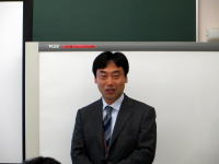 大平哲也氏がスーツを着て講義室で立っている写真。背景にはホワイトボードが見える。