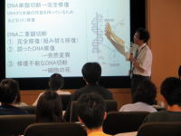 講義中の講師がDNAの構造について説明している様子。聴講者が数名座っている。