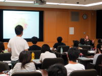 大学の教室で講義を受ける学生たちと、プレゼンテーションを行う講師の様子。