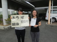 健康をテーマにした看板を持つ二人の女性が、福島県立医科大学の前で笑顔でポーズをとっている様子。