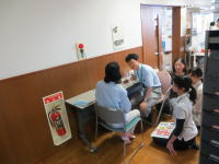 医療現場での学生たちが患者に対して診察を行っている様子。壁には消火器の表示が見える。