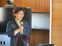 講演を行う女性がマイクを持ち、聴衆に向かって話している様子。背景にはテレビモニターと木製の壁が見える。