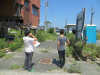 廃墟となった建物の前で、二人の男性が周囲を見渡している風景。青空が広がる中、雑草が生い茂る道が印象的。