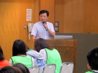 福島県立医科大学で講演を行う男性と、緑のベストを着た聴衆の様子。