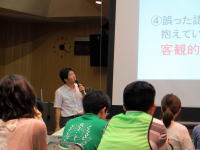 講演を行う男性と聴衆がいる会場の様子。スライドには「客観的」と書かれている。