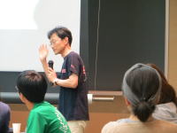 講演中の男性がマイクを持ち、聴衆に向かって話している様子。背景にはプレゼンテーション用のスクリーンが見える。