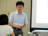 講義中の男性がプレゼンテーションを行い、受講者がメモを取っている様子。