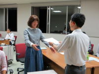 学生が会議室で書類を交換している様子。手前にはテーブルがあり、背景には窓と他の参加者が見える。
