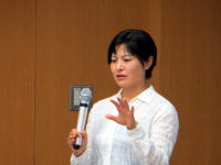 講演中の女性がマイクを持ち、手を挙げて話している様子。背景は木目調の壁。