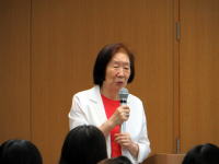 医療に関する講演を行う女性がマイクを持ち、聴衆に向かって話している様子。背景は木製の壁。