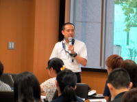 講演を行う男性がマイクを持ち、聴衆に向かって話している様子。背景には窓があり、参加者が集中している。
