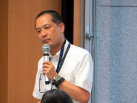 講演中の男性がマイクを持ち、聴衆に向かって話している様子。背景には窓が見える。