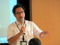 講演中の男性がマイクを持ち、聴衆に向かって話している様子。背景にはプレゼンテーションのスライドが映し出されている。