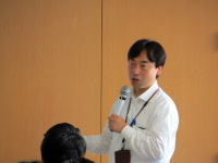 講演を行う男性がマイクを持ち、聴衆に向かって話している様子。背景は木目調の壁。