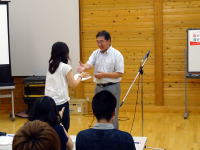 福島県立医科大学のイベントで、講師が参加者に賞を授与している様子。背景には木の壁とプロジェクターが見える。