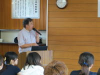 講演を行う男性がマイクを持ち、聴衆に向かって話している様子。背景には木製の壁と時計が見える。