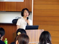 講演を行う女性がマイクを持ち、聴衆に向かって話している様子。背景には木の壁が見える。