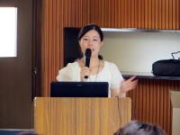 講演を行う女性がマイクを持ち、聴衆に向かって話している様子。背景には木目調の壁と窓が見える。