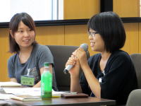 二人の女性が会議室で話し合いをしている様子。左側の女性は微笑み、右側の女性はマイクを持って発言している。