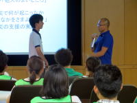 大学の講義中、講師が学生に話しかけている様子。聴衆は緑の服を着た学生たち。