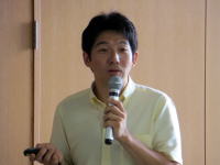 講演中の男性がマイクを持ち、聴衆に向かって話している様子。背景は木製の壁。
