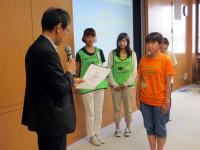 福島県立医科大学での表彰式の様子。緑のTシャツを着た学生たちが壇上で発表を聞いている。