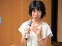 講演中の女性がマイクを持ち、聴衆に向かって話している様子。背景には木の壁が見える。