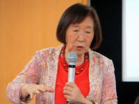 講演中の女性がマイクを持ち、熱心に話している様子。背景には木目調の壁が見える。