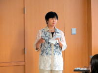 講演を行う女性が、木製の壁の前で話している様子。