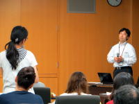 講演を行う男性と質問する女性がいる会議室の様子。参加者が聴講している。