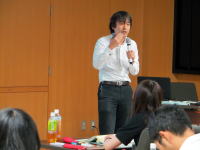 講演を行う男性がマイクを持ち、聴衆に向かって話している様子。背景には木製の壁とテーブルが見える。