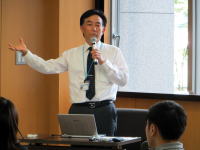 福島県立医科大学で講演を行う男性がマイクを持ち、聴衆に向かって話している様子。
