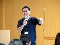 医療に関する講演を行う男性がマイクを持ち、聴衆に向かって話している様子。背景には木製の壁が見える。
