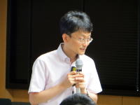 講演中の男性がマイクを持ち、聴衆に向かって話している様子。背景には窓があり、明るい室内の雰囲気が感じられる。