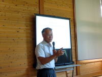 講演を行う男性がマイクを持ち、木製の壁の前で話している様子。背景にはスクリーンが設置されている。