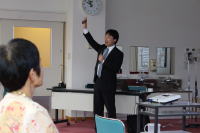 講演を行う男性が手を挙げている様子。聴衆には女性が座っており、医療に関するセミナーの一場面。