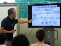 災害医療に関する講義で、講師がスライドを指し示しながら説明している様子。受講者が後方に座っている。