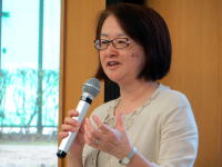 講演中の女性がマイクを持ち、手を使って話している様子。背景には窓があり、自然光が差し込んでいる。