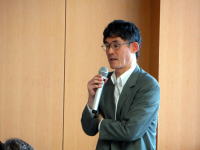 講演中の男性がマイクを持ち、聴衆に向かって話している様子。背景は木製の壁。