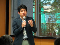 臨床心理士がマイクを持ち、福島の避難者の心理について講演している様子。背景には窓からの緑が見える。